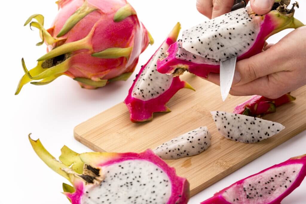 Closeup of pitaya fruit flesh being removed from its magenta colored skin. A cook’s left hand holding a fruit wedge, while his right is cutting with a kitchen knife. Differential focus. White ground.