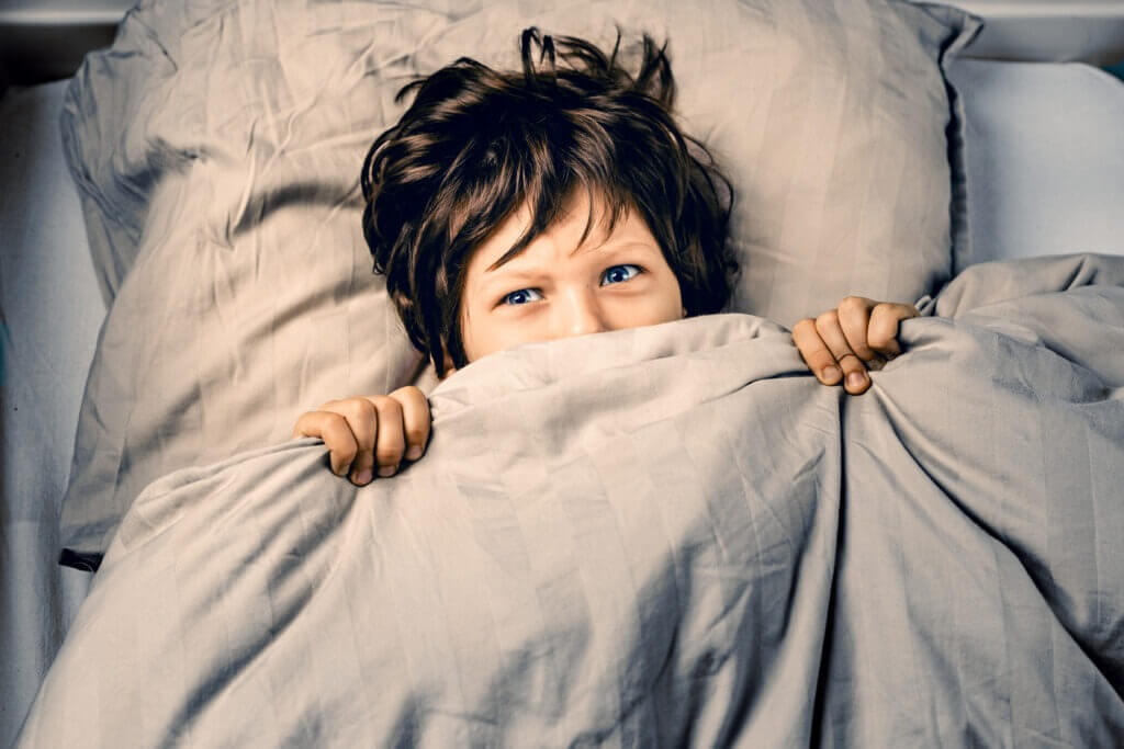 Little boy covering his face with a blanket is about to cry being scared of nightmares, ghosts or monsters in his bedroom. 