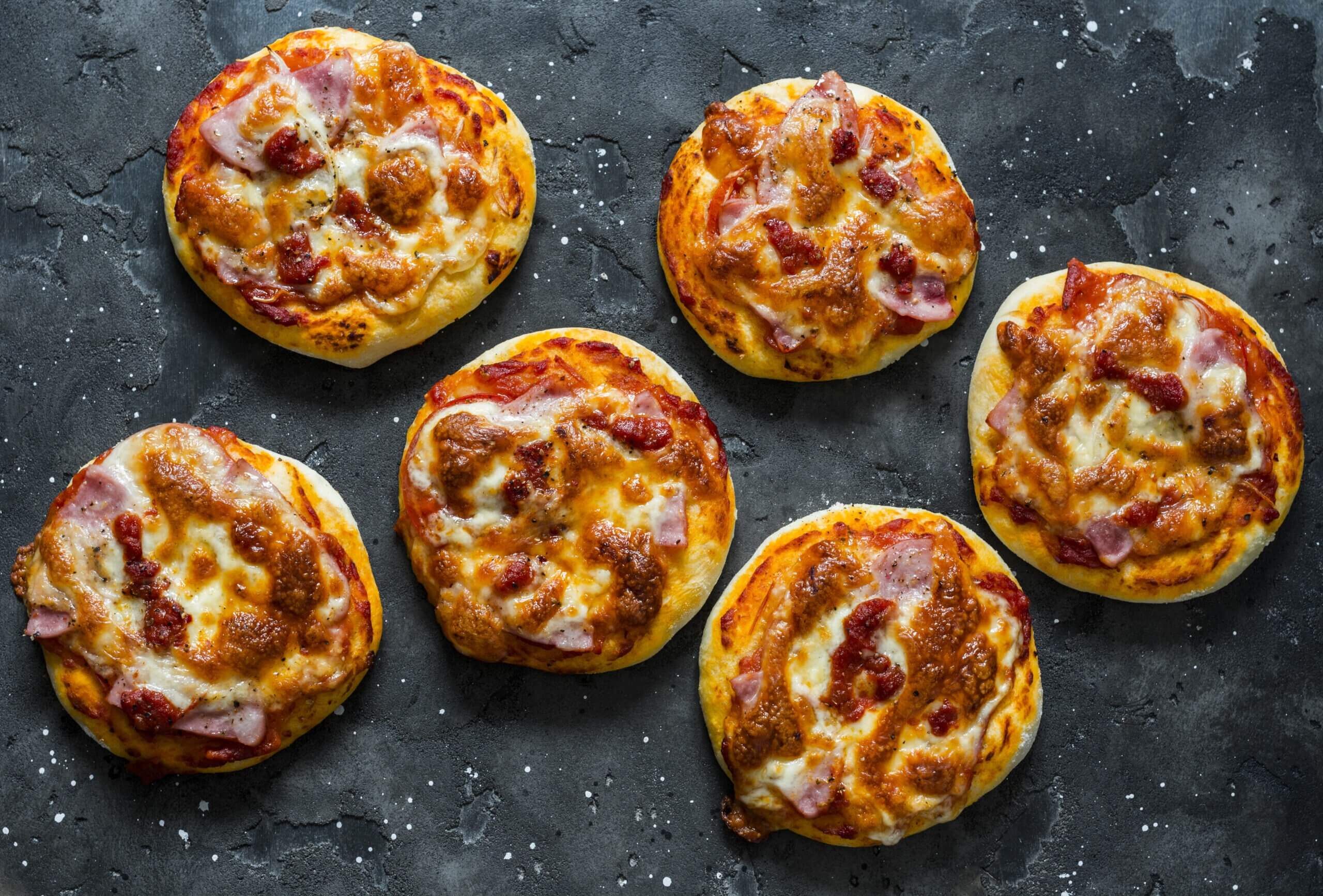 Mini ham, tomato sauce, mozzarella, sun dried tomatoes pizza on a dark background, top view.