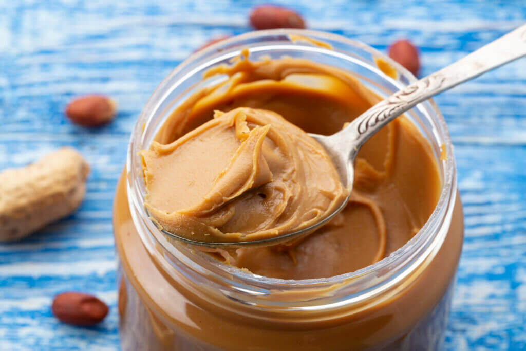 Peanut butter in an open jar and peanuts in the skin are scattered on the blue table.