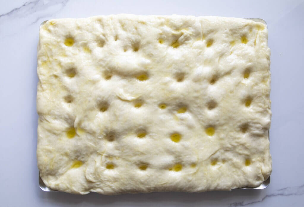 Traditional focaccia dough made from flour, water, yeast, sugar, salt water, and olive oil holes - top view, cooking process on a baking sheet on a marble table