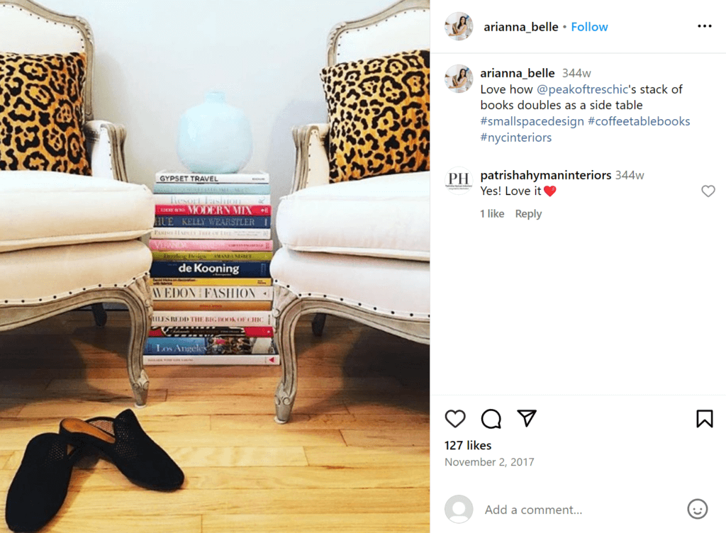 Two white chairs with leopard print throw pillows; in between is a stack of fashion books used as an end table with a blue round vase on top.