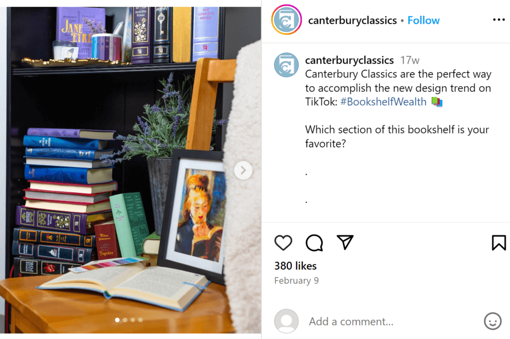 Black bookshelf featuring mainly blue and purple books, with a wooden chair in front acting as a shelf for a small framed painting and a book that is laying flat open. 