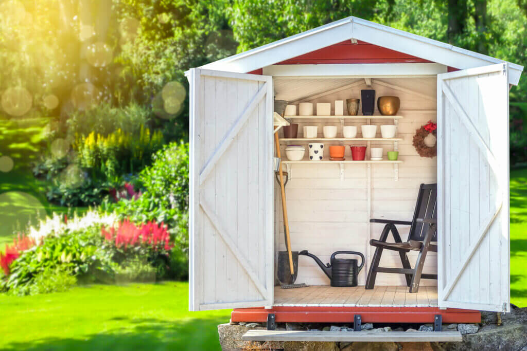 Garden shed filled with gardening tools. Shovels, rake, pots, water pitcher in storage hut. Green sunny garden in the background.