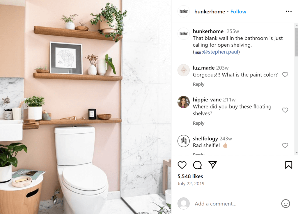 Open shelving above a toilet on a blush pink wall in a bathroom, featuring several plants and a framed photo