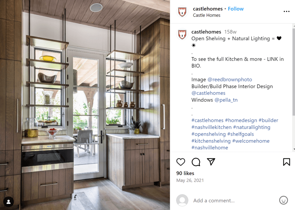 Modern open shelves hanging from the ceiling in front of the windows, which look out onto a patio. Shelves contain kitchen items like bowls and pots.