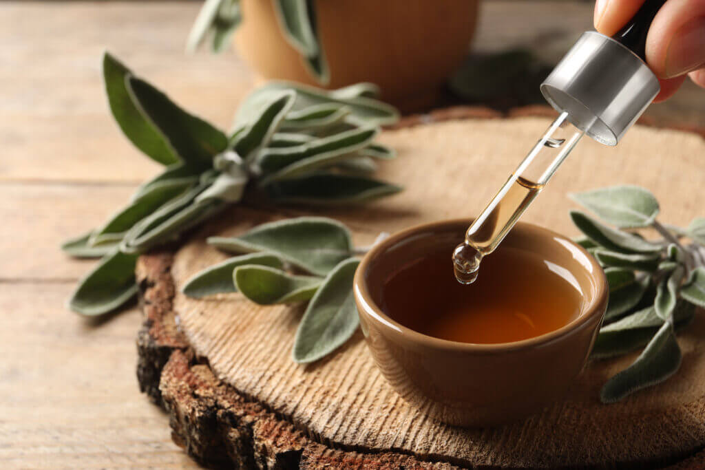 Woman dropping essential sage oil into bowl at wooden table, closeup. 