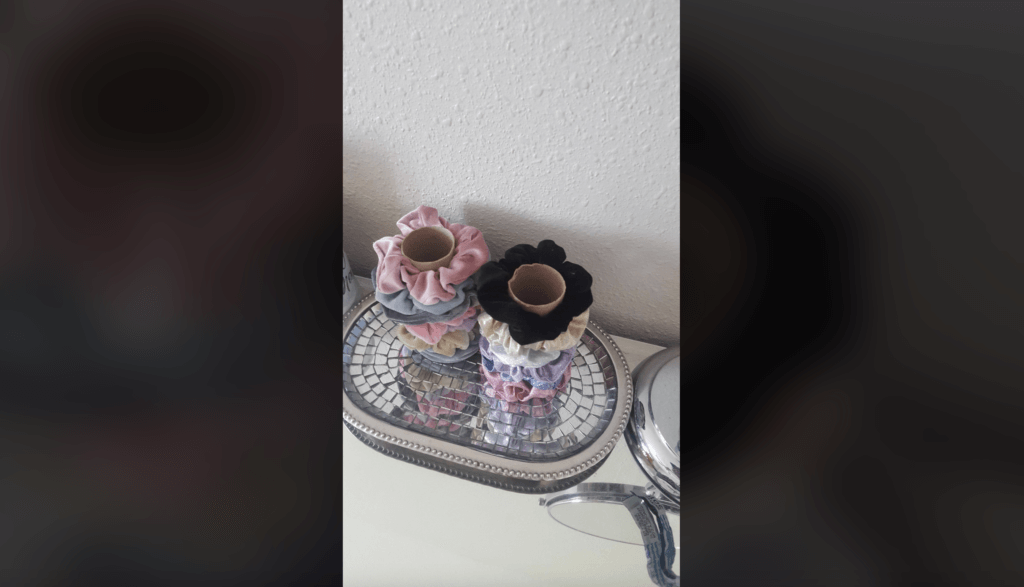 A mirrored tray holds two cardboard tubes stacked with colorful scrunchies in shades of pink, black, beige, blue, and more. The tray is placed on a white surface next to a small round mirror, reflecting common household items used for hair styling.