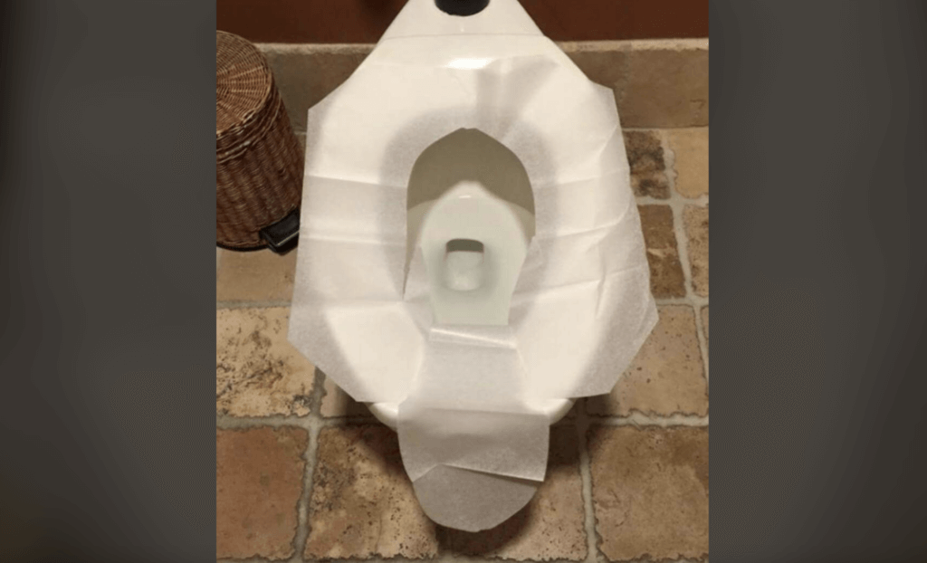 Image showing a toilet with multiple layers of toilet seat covers carefully arranged to cover the entire seat. The floor features beige tiles, and a wicker basket is visible to the left