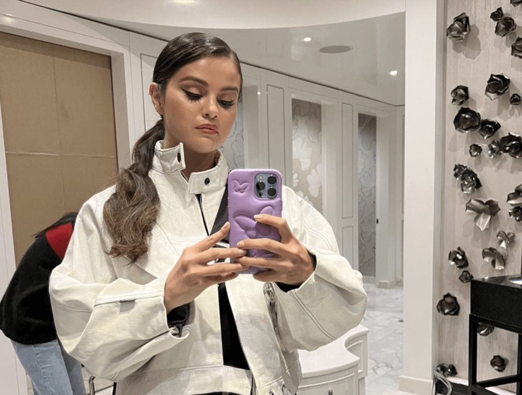 Selena Gomez in a white jacket taking a mirror selfie