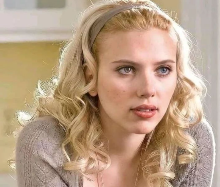 Scarlett Johansson is shown with blonde, curly hair, styled with a grey headband. She wears a light grey sweater and has a thoughtful expression, her blue eyes looking off-camera. The background is softly lit, adding a warm and calm atmosphere to the scene.