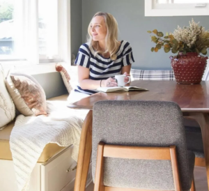 A woman sits at a cozy breakfast nook with sunlight streaming through the window, holding a mug and an open book. She wears a striped shirt and looks outside, next to a table decorated with a red vase filled with dried flowers. The space is accented with pillows and a blanket, creating a warm, inviting atmosphere.