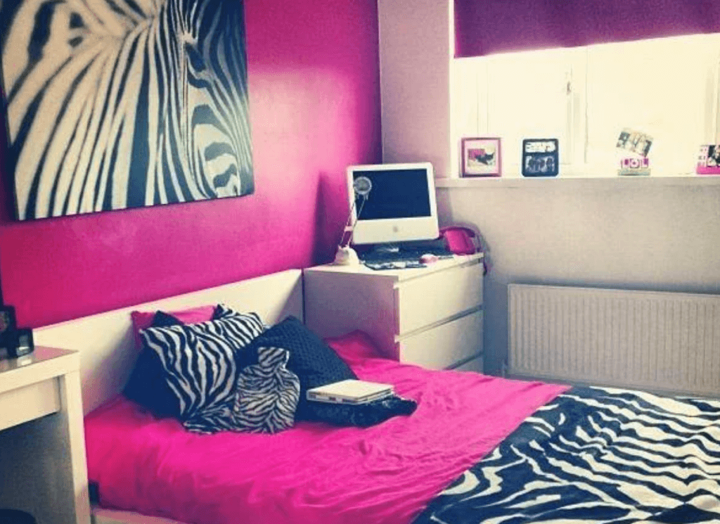 A brightly decorated bedroom with a pink and zebra print theme. The bed features pink bedding with zebra print pillows and a matching zebra print blanket. On the wall above the bed is a large zebra print artwork, and there is a desk with an iMac computer and some framed photos on the windowsill.