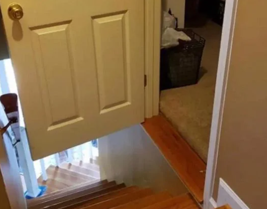 A door that opens directly over a staircase, with the bottom edge of the door cut to fit above the steps. The setup appears hazardous, as it does not provide safe passage from the carpeted room to the wooden stairs below.