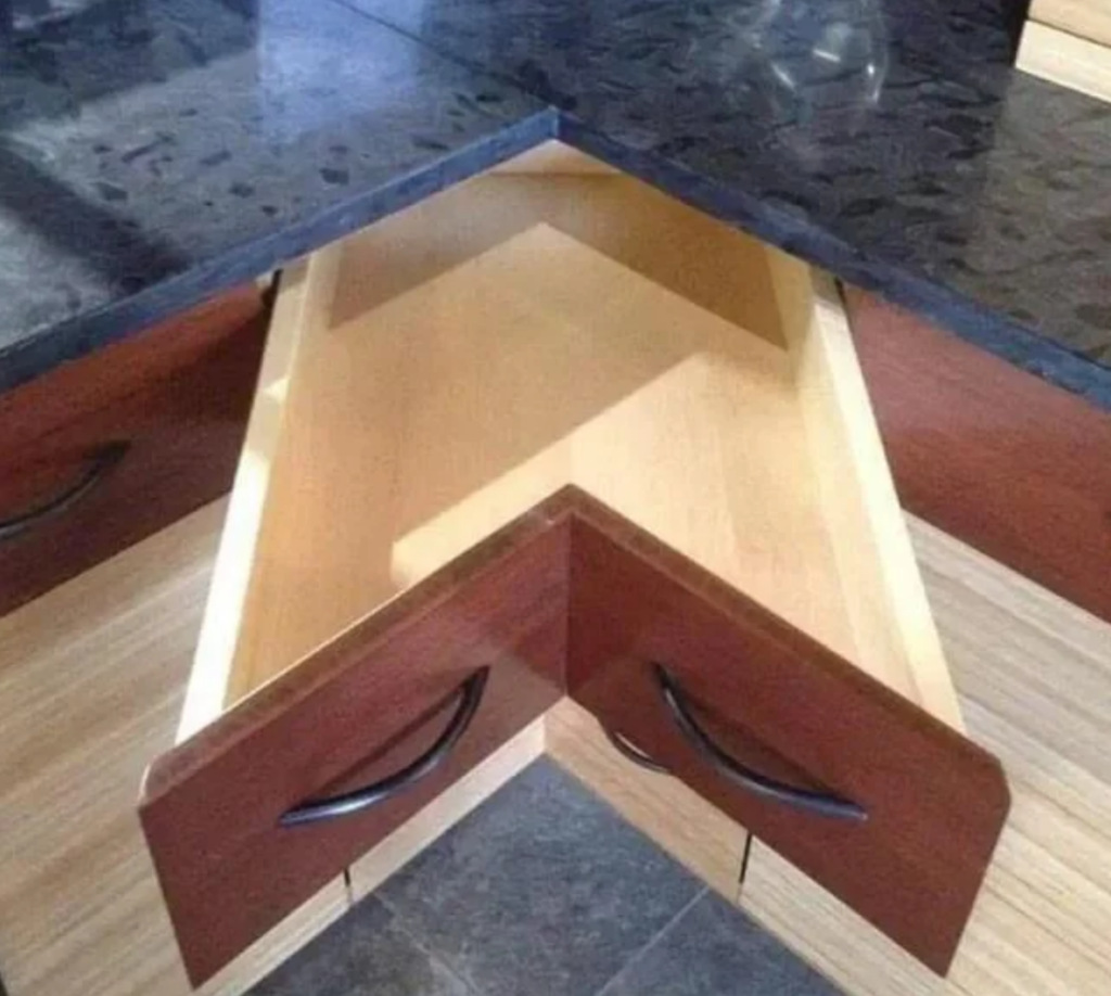 A kitchen drawer designed to fit into a corner, resulting in a triangular shape that pulls out at an angle. The drawer has two handles, but its awkward shape and placement make it impractical for regular use.