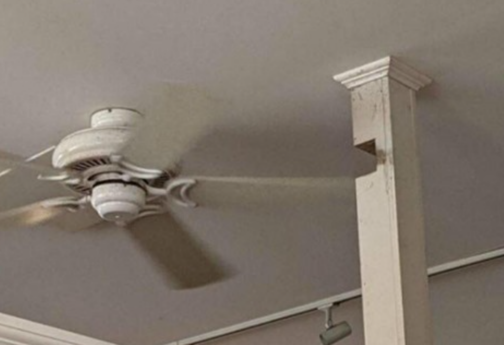 A ceiling fan with its blades obstructed by a wooden support column, making the fan unusable. The setup suggests a significant design flaw, as the fan's functionality is compromised by the poorly placed column.