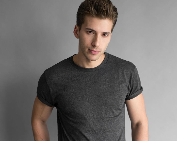 Scarlett Johansson's twin brother, Hunter Johansson, stands against a plain gray background wearing a dark gray t-shirt. He has short, styled hair and is looking directly at the camera with a slight smile.