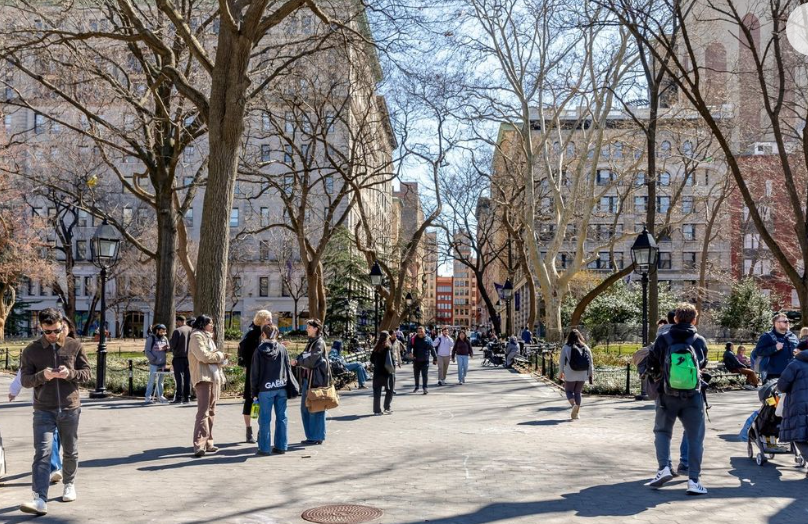 Students and pedestrians walk through a tree-lined pathway on the NYU campus. The scene is bustling with activity, set against a backdrop of historic buildings and clear, sunny skies.