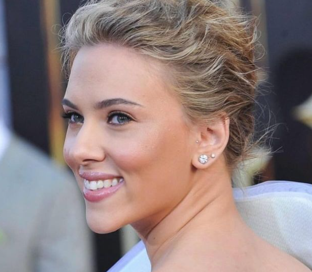 Scarlett Johansson, with her blonde hair styled in an elegant updo, smiles warmly over her shoulder, showcasing her sparkling earrings and subtle makeup. The softly blurred background highlights her glowing complexion and radiant expression.