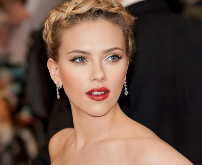 Scarlett Johansson is pictured with a sophisticated braided updo, striking red lipstick, and elegant dangling earrings. Her makeup is flawlessly applied, emphasizing her blue eyes and creating a glamorous look against a blurred background.