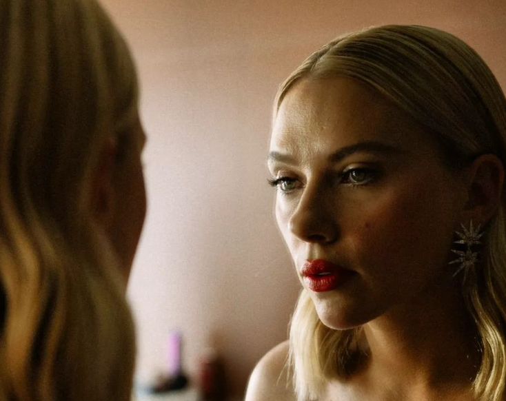 Scarlett Johansson is seen in profile, looking into a mirror with a contemplative expression. She wears striking red lipstick and starburst-shaped earrings, her blonde hair styled straight. The soft lighting creates a warm, intimate atmosphere.