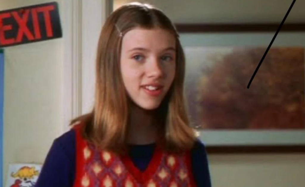 A young Scarlett Johansson is seen in a scene from "Home Alone 3." She has straight, shoulder-length hair with a hair clip, and she wears a red and orange patterned vest over a blue shirt. A sign reading "EXIT" is visible in the background, adding to the familiar setting of the movie.