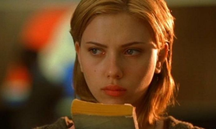 Scarlett Johansson, with a serious and contemplative expression, is holding a yellow book. She has shoulder-length blonde hair, and the soft lighting creates a warm, introspective atmosphere. The blurred background features muted colors, keeping the focus on her face.