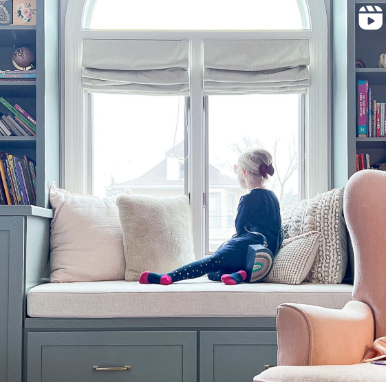 A young child sits on a cozy window seat, surrounded by plush cushions, gazing out the window at a peaceful neighborhood scene. The window seat is flanked by built-in shelves filled with books, creating a warm and inviting reading nook.