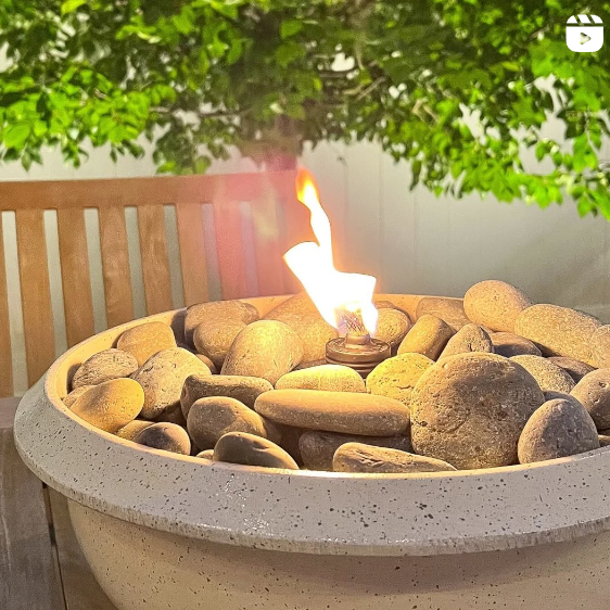 A small fire burns brightly in a tabletop fire pit filled with smooth stones, set against a backdrop of lush greenery and a wooden chair. The warm, inviting scene suggests an outdoor gathering or a cozy evening on a patio.