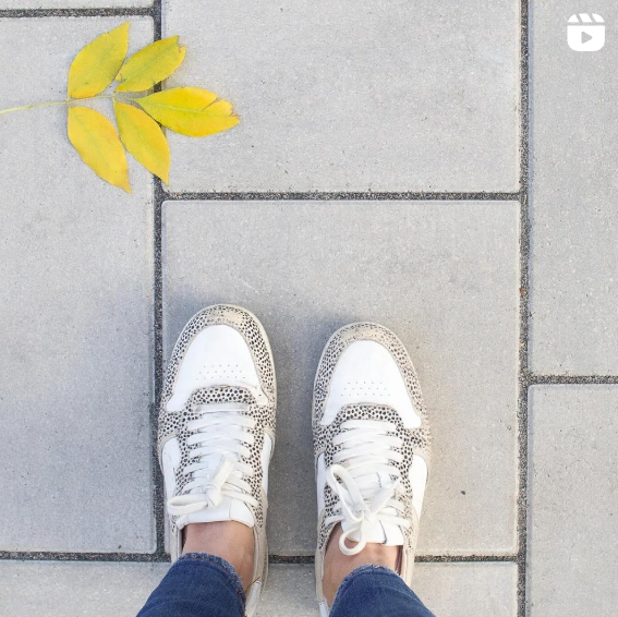 A pair of white sneakers with black speckles stand on a neatly arranged stone pavement, next to a fallen yellow leaf. The scene captures a casual moment of autumn, emphasizing the change of seasons.