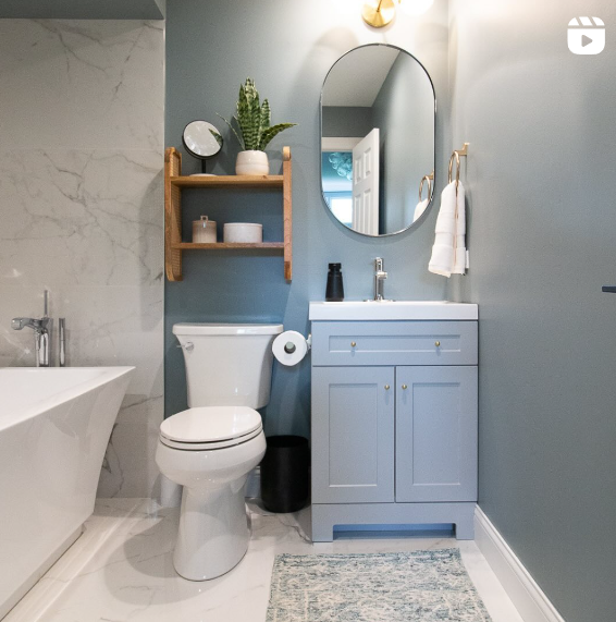 A modern bathroom features a freestanding bathtub, a toilet, and a blue vanity with a round mirror above it. The walls are painted a soothing grayish-blue, complemented by white marble accents and a small wooden shelf holding decorative items and a plant. The space is well-lit, with a cozy and elegant atmosphere.