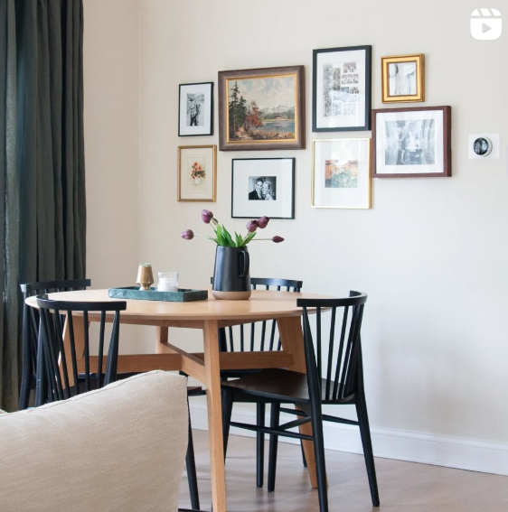 A cozy dining area features a round wooden table with black chairs, a vase of tulips, and a gallery wall filled with various framed artworks and photographs. The space is bright and inviting, with a curtain to one side and a neutral color scheme that highlights the eclectic mix of wall art.