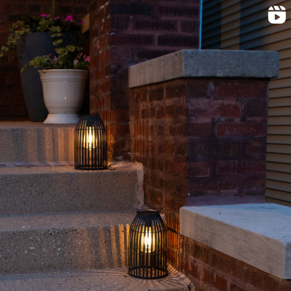 An outdoor stairway at dusk is illuminated by two black lanterns with glowing lights inside, creating a warm and inviting atmosphere. The steps are flanked by brick walls, and a potted plant adds a touch of greenery to the scene.