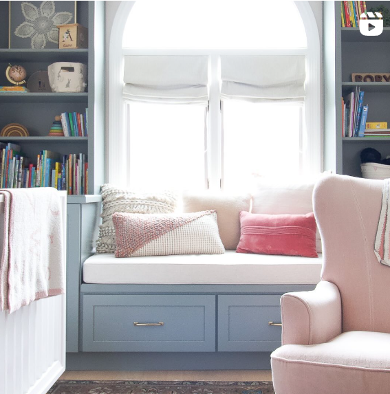 A cozy window seat adorned with various cushions in neutral and pink hues is set against a large, arched window. The surrounding built-in shelves are filled with books and decorative items, creating a warm and inviting reading nook. A pink armchair sits nearby, enhancing the comfortable atmosphere.