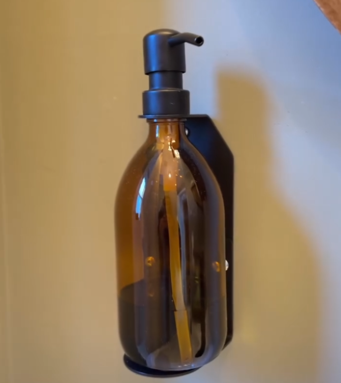 A wall-mounted amber glass soap dispenser with a black pump and holder is installed against a beige wall. The sleek and minimalist design adds a touch of elegance and functionality to the space.