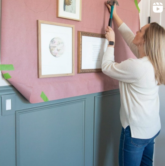 A woman is hanging framed art on a wall using a sheet of paper as a guide for placement. The wall features a two-tone design with a blue-gray wainscoting and white upper section, adding a decorative touch to the space. She is carefully hammering nails into the wall to ensure precise alignment of the frames.