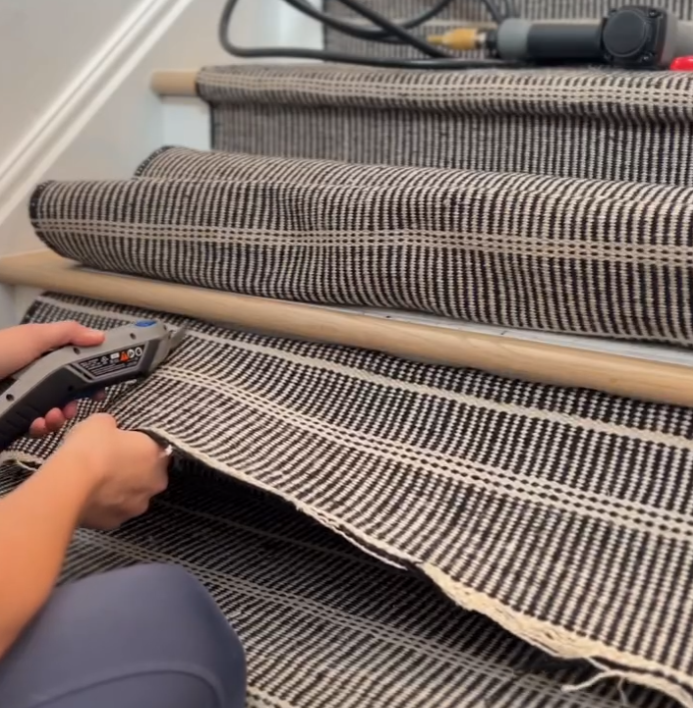 A person is installing a striped runner on a staircase, using a staple gun to secure the fabric. The runner is being carefully laid out and attached to each step, ensuring a neat and snug fit. Tools and materials are visible on the stairs, indicating an ongoing home improvement project.