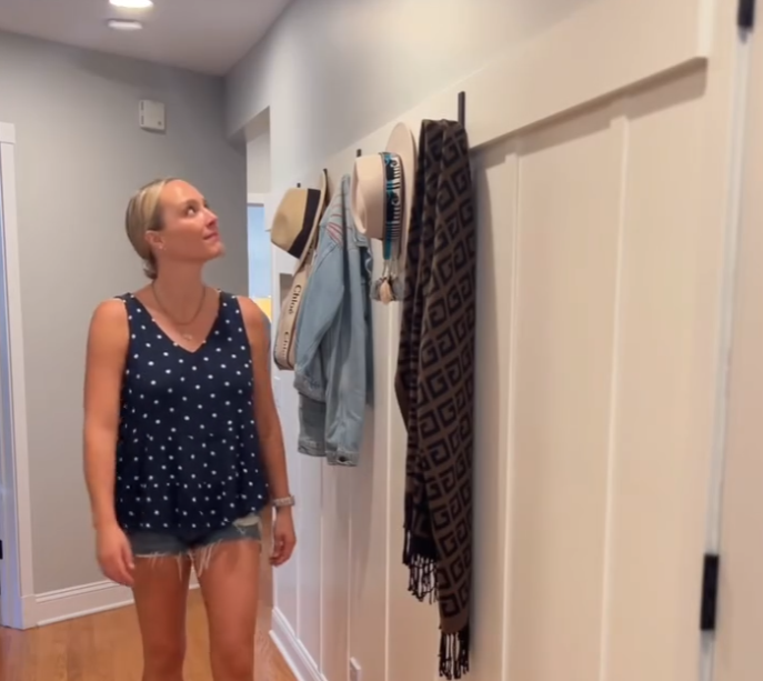 A woman in a polka-dot sleeveless top and denim shorts admires a wall-mounted coat rack in a hallway. The rack holds various items, including hats, a denim jacket, and a patterned scarf, showcasing a stylish and organized entryway.