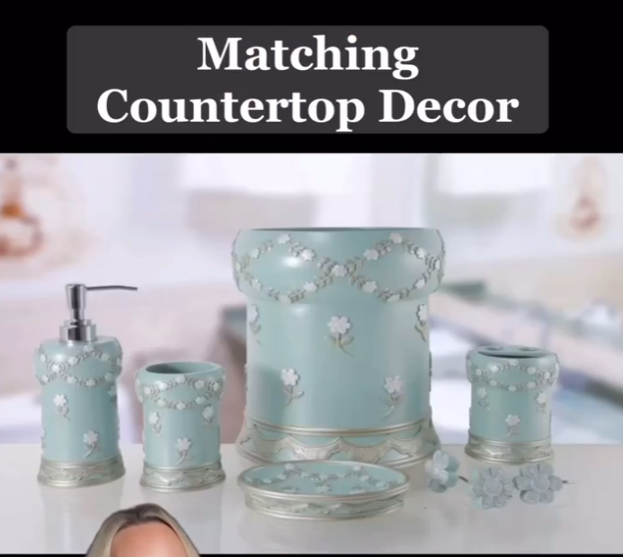 A set of matching countertop decor items is displayed, featuring a soap dispenser, toothbrush holder, tumbler, waste bin, and a soap dish. The items are in a pastel blue color with delicate floral and lace-like patterns, creating an elegant and coordinated look for a bathroom. The text overlay reads, "Matching Countertop Decor."