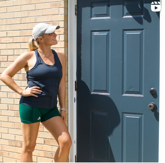 A woman dressed in a navy tank top, green shorts, and a gray cap stands next to a dark blue front door, leaning against a brick wall. She appears to be enjoying a sunny day, possibly after a workout or run.