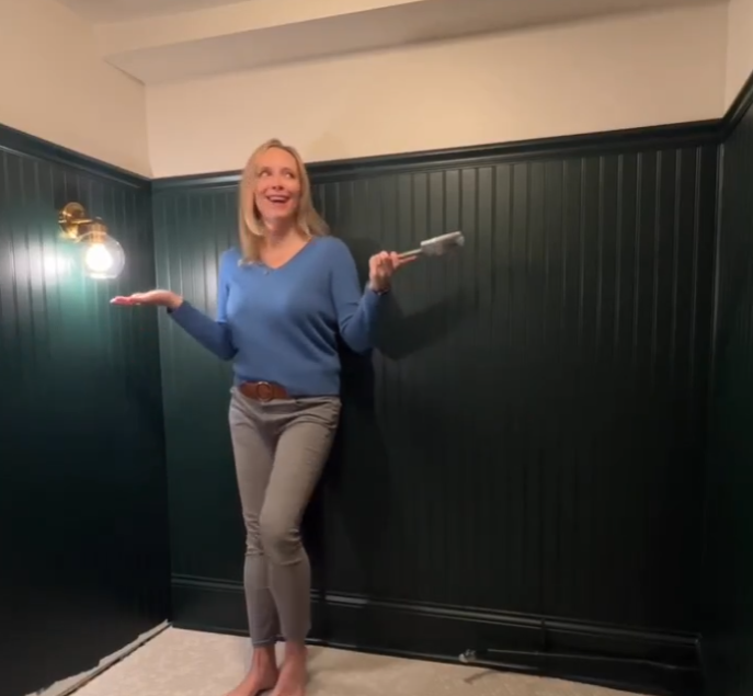 A woman in a blue sweater and gray pants stands in a room with dark green wainscoting and cream-colored upper walls, holding a paint roller. She appears cheerful and satisfied, suggesting she is in the process of or has just completed a painting project. A brass light fixture illuminates the space, adding a warm touch to the freshly updated room.