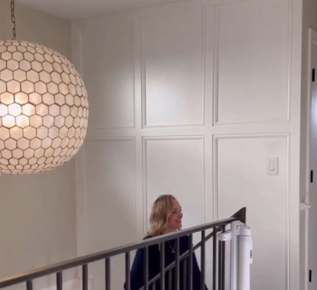 A woman stands at the bottom of a staircase, smiling and looking up. The staircase is adjacent to a wall with elegant white paneling, and a large, spherical light fixture with a honeycomb pattern hangs above. The space exudes a modern and sophisticated ambiance.