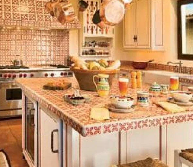 A warm, inviting kitchen featuring a large island with a decorative tile countertop, styled in a 90s fashion. The island is set for breakfast with bowls, mugs, and glasses of juice. Hanging above the island are copper pots and pans, and the backsplash is adorned with intricate tile work. The kitchen also has a stove, various cooking utensils, and a basket of fresh bread and fruits on the island.