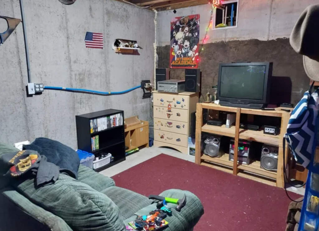 A basement room with a nostalgic 90s and 2000s vibe, featuring a CRT television, a GameCube console, and a stereo system on a wooden shelf. The room has a green couch, a dresser with stickers, a small American flag on the wall, and a "My Hero Academia" poster. The setup includes shelves with video games and DVDs, and the space is lit with colorful string lights.