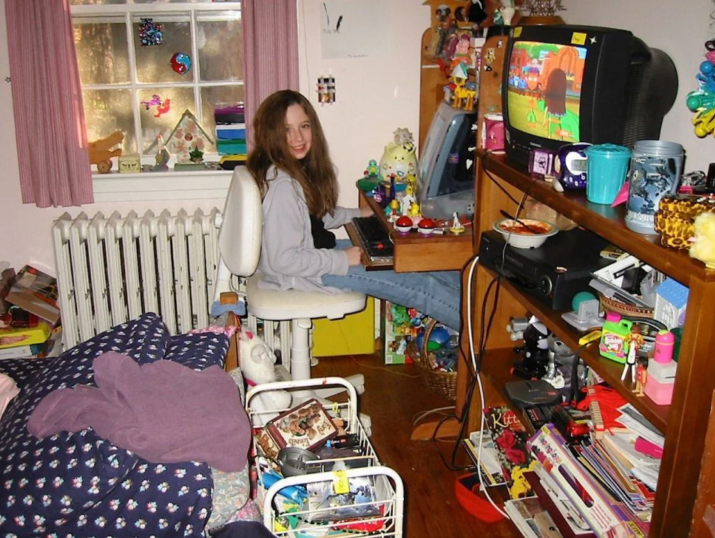 A cozy bedroom from the 90s or 2000s featuring a girl sitting at a desk with a computer and a CRT television displaying a cartoon. The room is filled with colorful decorations, toys, and collectibles on shelves. There's a bed with patterned bedding, a radiator under the window, and a cart with VHS tapes and other items. The girl is smiling, surrounded by the nostalgic charm of the era.