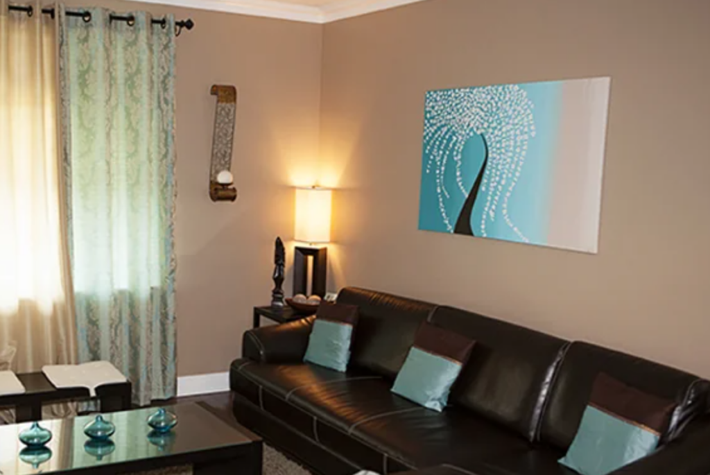 A modern living room with a 2000s aesthetic, featuring a dark brown leather couch adorned with light blue and brown pillows. A contemporary painting of a tree in blue and white tones hangs on the wall above the couch. The room is softly lit by a floor lamp next to the couch, and light filters in through patterned curtains. A glass coffee table in front of the couch holds decorative blue glass pieces.