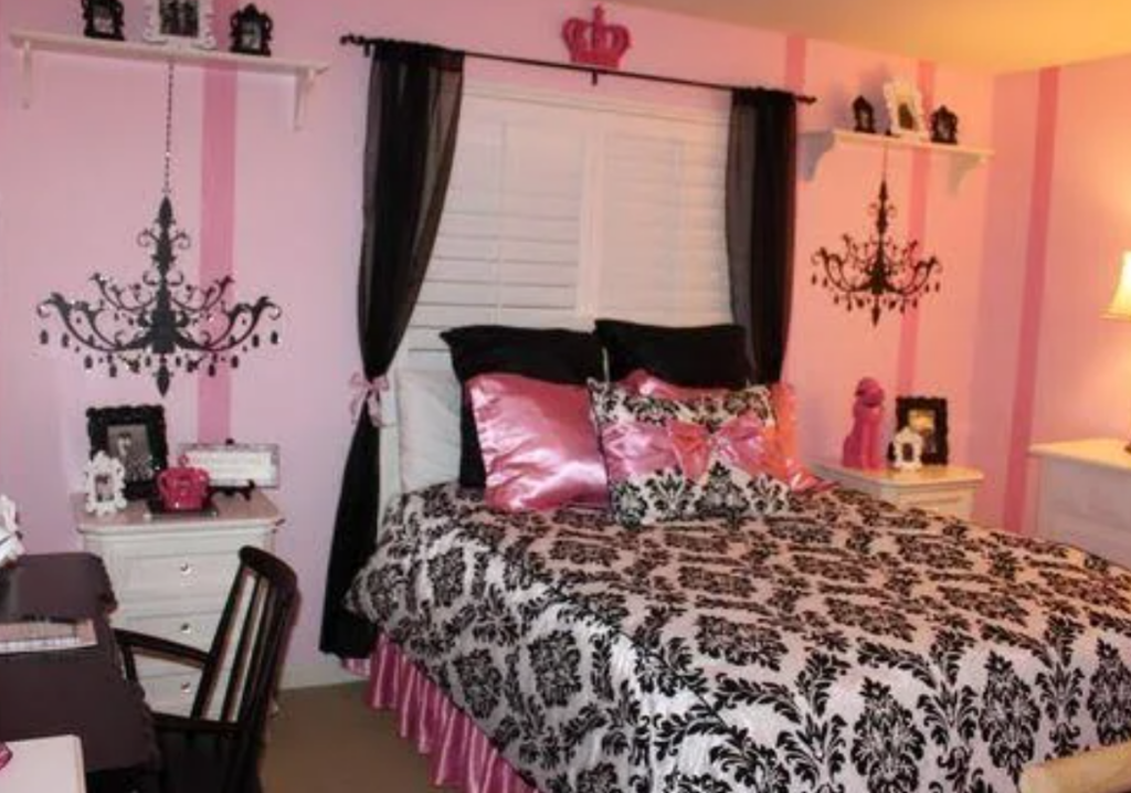 A stylish bedroom with a pink and black color scheme, typical of the 2000s. The bed features black and white damask patterned bedding, complemented by pink satin pillows. The walls are decorated with black chandelier decals, and the room includes white furniture such as a nightstand and desk. Decorative items like framed photos, a pink poodle statue, and a crown motif add to the chic, feminine aesthetic.