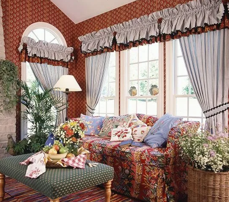 A living room decorated in a 90s and 2000s style, featuring a floral-patterned couch with an array of colorful cushions. The room has large windows with elaborate blue and white curtains adorned with ruffled valances. A green upholstered ottoman serves as a coffee table, holding a fruit basket and a vase of flowers. The room is further embellished with patterned wallpaper, a floor lamp, houseplants, and a basket of white flowers, creating a cozy and vibrant atmosphere.