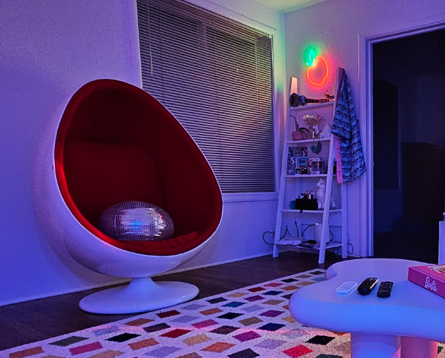 A modern living room with a retro-futuristic feel, featuring a red egg chair with a disco ball pillow, evoking a style from the 2000s. The room has a colorful, geometric-patterned rug, a white coffee table with remotes and a "Barbie" game, and a shelf decorated with various knick-knacks and a small plant. Neon lights in red and green add a vibrant touch, and a window with blinds completes the cozy yet stylish ambiance.