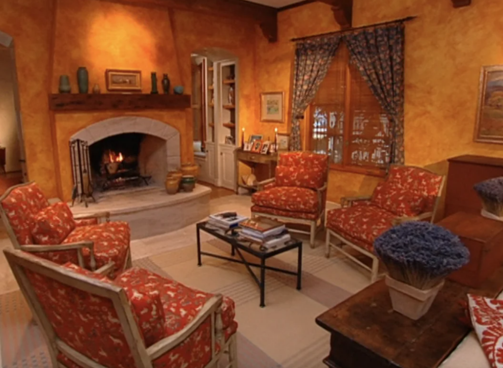 A cozy living room with a rustic 90s or 2000s design, featuring warm yellow and orange textured walls and a stone fireplace. The room includes four armchairs with red and white patterned upholstery arranged around a small coffee table. The mantel above the fireplace is adorned with decorative vases, and framed pictures are displayed on a side table. Floral curtains frame the windows, and potted lavender plants add a touch of greenery to the inviting space.